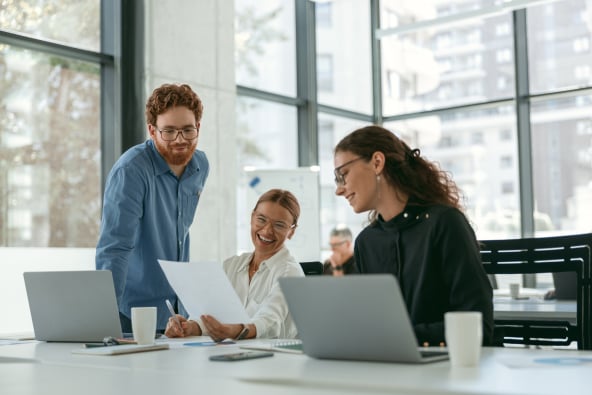 A group of colleagues in a sunny office chat and laugh over work at a conference table with laptops