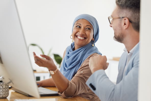 Two colleagues smile and collaborate seated at a sunny table with a laptop