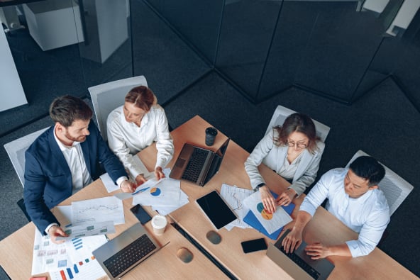 A group of sharply dressed professionals seated at a conference table with laptops and documents