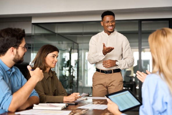 A group of colleagues converse at a conference table and one person is standing to present