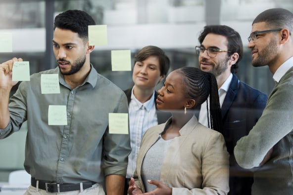 A group of professionally dressed people look at sticky notes posted to a glass wall and collaborate