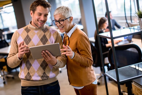 Two colleagues smile and converse looking at a tablet in an office space